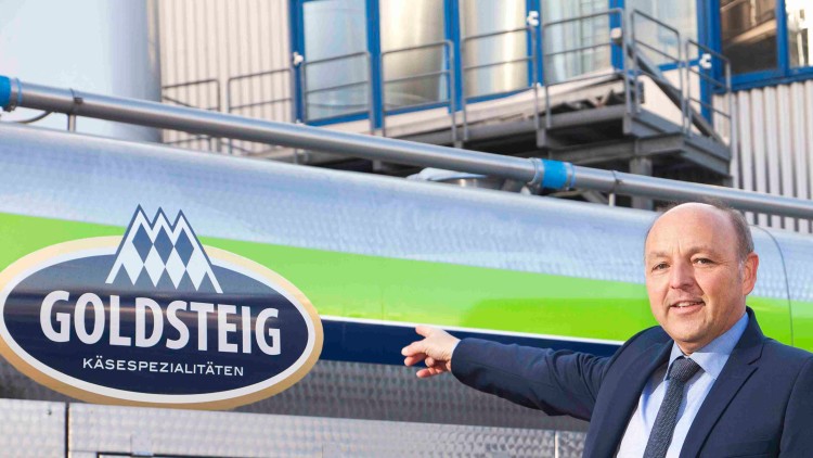 Ein Mann im Anzug zeigt auf ein silbernes Fahrzeug mit dem Logo „GOLDSTEIG Käsespezialitäten“, davor ein gelbes Schild mit „Gutes von hier“.