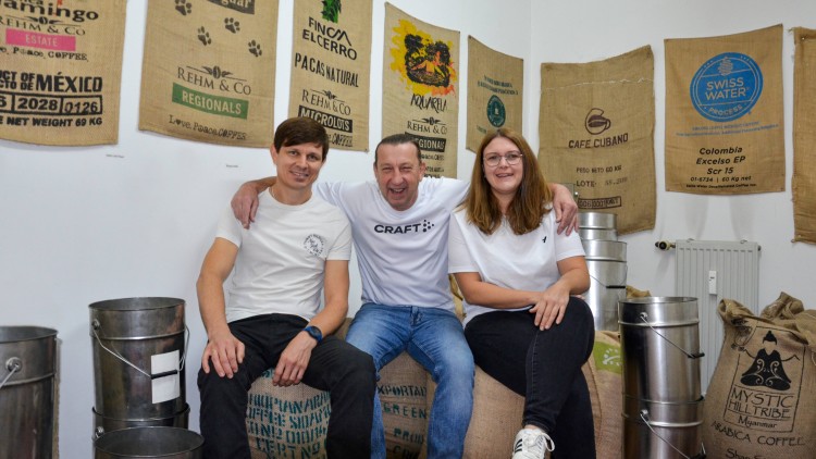 Drei Personen sitzen auf Kaffeesäcken vor einer Wand mit bedruckten Jutesäcken und neben metallenen Behältern.