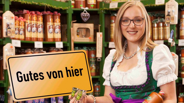 Frau in Tracht hält regionale Produkte in der Hand, im Hintergrund Regale mit Lebensmitteln, Schild mit Text: Gutes von hier.