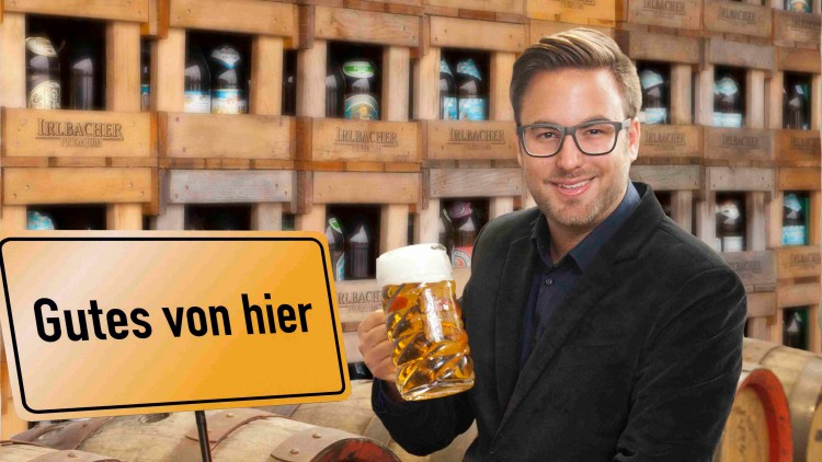 Ein Schild mit der Aufschrift „Gutes von hier“ steht neben Bierflaschen, einem Bierkrug und Holzfässern aufgestapelter Bierkisten.