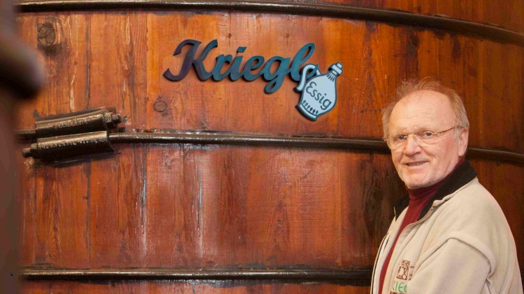 Person vor einem großen hölzernen Essigfass mit der Aufschrift „Krieg Essig“ und einem gelben Schild „Gutes von hier“.