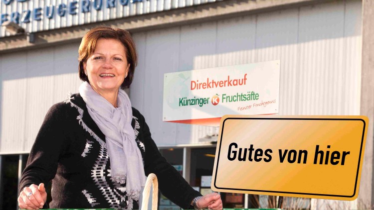 Ein Korb mit Äpfeln und Flaschen der Marke Künzinger Fruchtsäfte steht vor Kisten, ein Schild mit „Gutes von hier“ ist zu sehen.