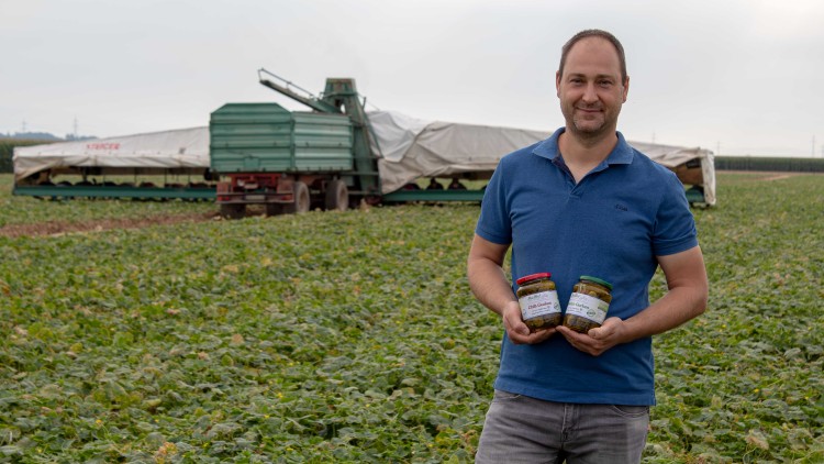 Eine Person steht auf einem grünen Feld vor einem Erntefahrzeug und hält zwei Gläser mit beschriftetem Etikett in den Händen.