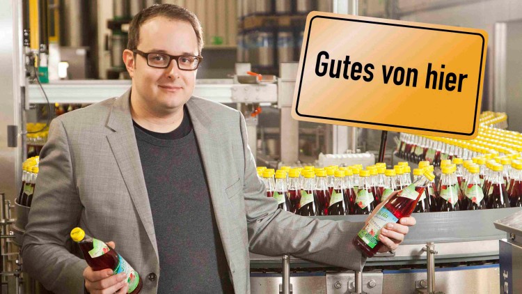 Person hält zwei Glasflaschen mit gelbem Deckel vor einer Abfüllanlage, Schild mit der Aufschrift Gutes von hier.