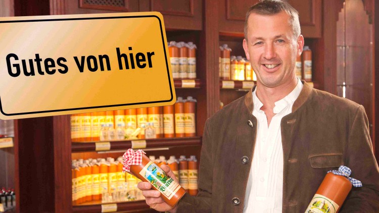 Person in Tracht hält zwei Flaschen Senf, im Hintergrund Regale mit Produkten, Schild mit der Aufschrift Gutes von hier.