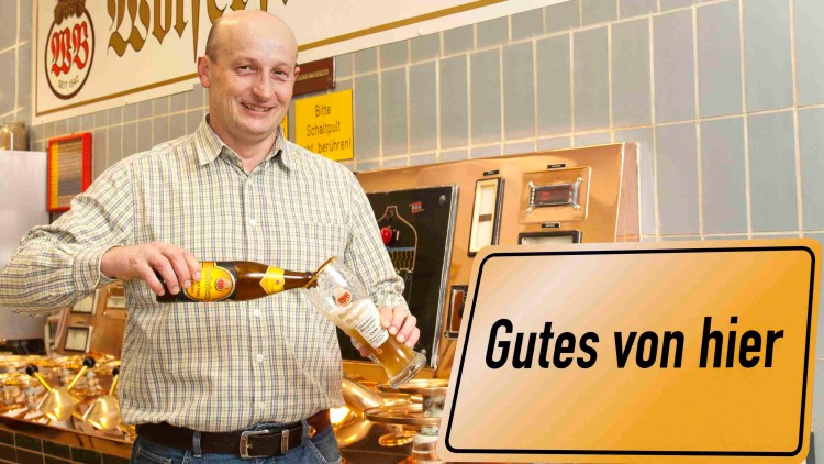 Person füllt Bier aus einer Flasche in ein Glas, im Hintergrund Schriftzug Wolferstetter Bier und Schild Gutes von hier.