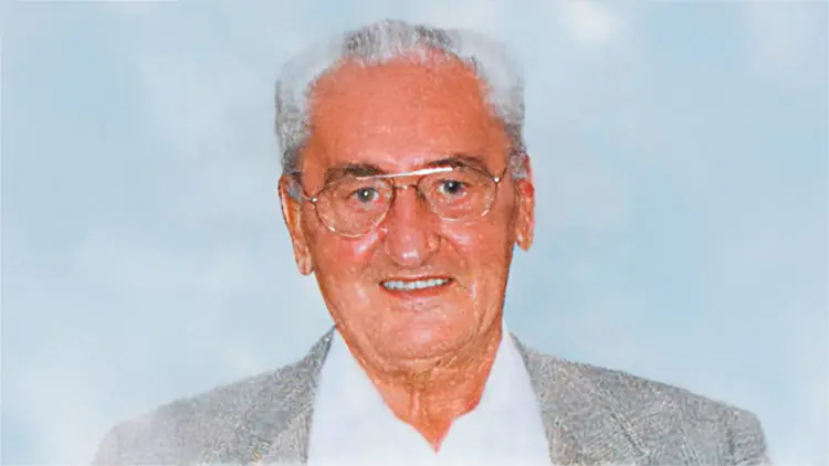 Eine ältere Person mit grauem Haar, Brille und hellem Sakko vor einem hellblauen Hintergrund.