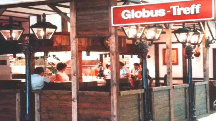 Der Globus-Treff mit Holzwänden und Laternen ist mit Menschen besetzt, das rote Schild mit weißer Schrift zeigt den Namen.