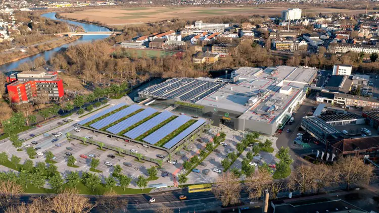 Ein GLOBUS Markt mit Parkplätzen und Solarpaneelen auf den Überdachungen, umgeben von Bäumen, neben einer Stadtlandschaft.