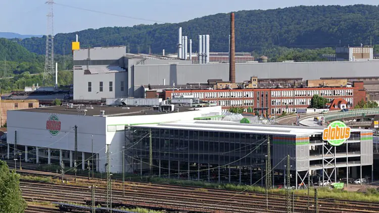 Das GLOBUS Gebäude mit Glaskonstruktion steht nahe einem Bahnhof, im Hintergrund befinden sich Industrieanlagen und bewaldete Hügel.