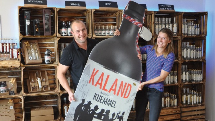 Zwei Personen halten eine große Nachbildung einer Flasche mit der Aufschrift „KALAND, MANGSHER KÜMMEL“ vor Holzregalen.
