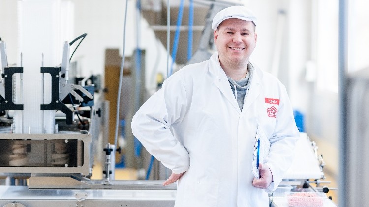 Person in weißem Arbeitskittel mit rotem Logo steht in einem hellen Industrie-Raum neben einer Metallmaschine.