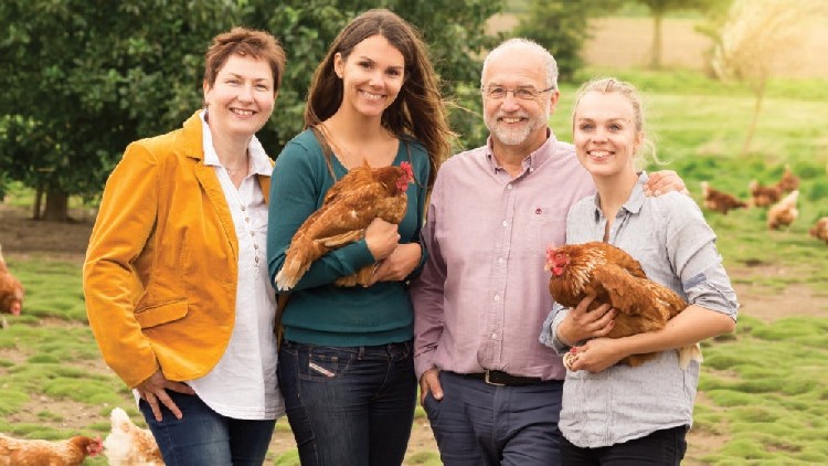 Vier Personen stehen auf einer grünen Wiese mit freilaufenden Hühnern und halten jeweils ein Huhn im Arm.