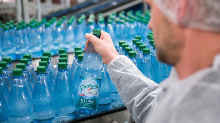 Eine Hand in weißem Ärmel hält eine Glasflasche „Glashäger Medium“, umgeben von weiteren Flaschen auf einem Fließband.