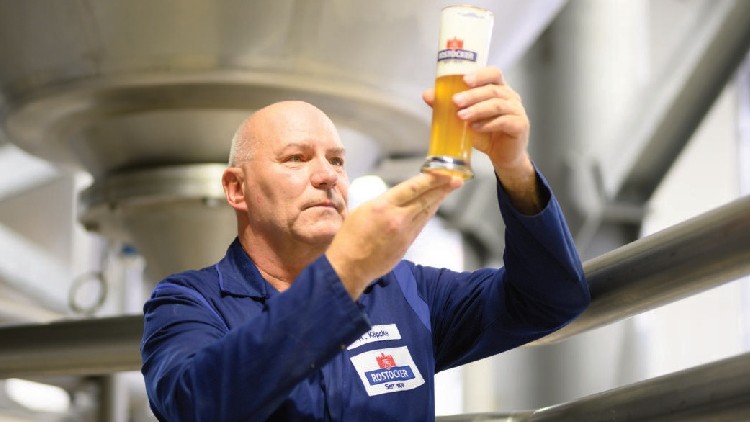 Person in blauer Arbeitskleidung hält ein Rostocker-Bierglas mit Schaumkrone vor metallischer Brauereikulisse hoch.