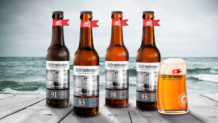 Vier Störtebeker-Bierflaschen und ein Störtebeker-Glas mit Bier stehen auf einem Holztisch vor einer Meereskulisse.