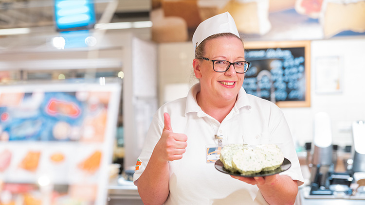 Eine Person in weißer Arbeitskleidung hält ein Stück Käse auf einem Teller und zeigt mit der anderen Hand einen Daumen hoch.