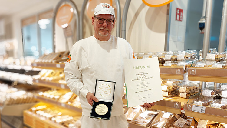 Eine Person in weißer Bäckerbekleidung hält eine Urkunde und ein schwarzes Etui mit einer goldenen Auszeichnung.