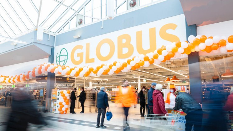 Eingang zu einem GLOBUS Markt mit orange-weißer Luftballon-Dekoration und einkaufenden Personen unter Glasdach.
