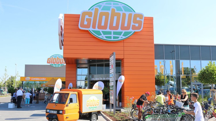 Orangefarbene GLOBUS-Fassade mit großem Logo, Getränkecenter, orangem Lieferwagen und Menschen bei Fahrrädern und Einkaufswagen.