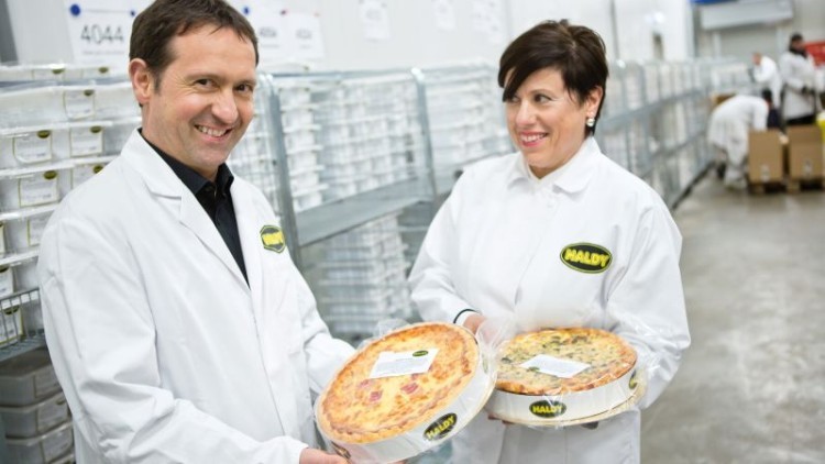Zwei Personen in weißen Arbeitskitteln halten runde Fertigpizzen mit HALDY-Logo in einer Produktionshalle.