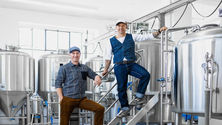 Zwei Personen stehen in einer Brauerei mit großen Edelstahlbehältern und industriellen Rohren im Hintergrund.