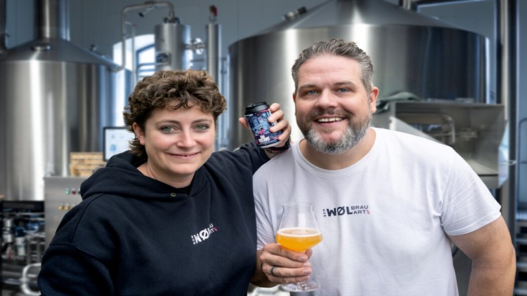 Zwei Personen in Brauerei tragen Kleidung mit „Wol Brau Art“-Logo, halten Getränkedose und Bierglas vor Chromtanks.