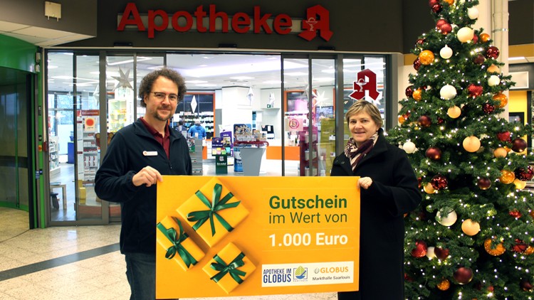 Zwei Personen halten einen gelben Gutschein mit der Aufschrift „Gutschein im Wert von 1.000 Euro“, daneben ein Weihnachtsbaum.
