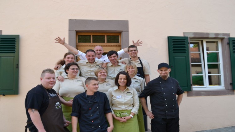 Eine Gruppe von Personen in Arbeitskleidung steht vor einem Gebäude mit hellbeiger Wand, grünen Fensterläden und zwei Fenstern.