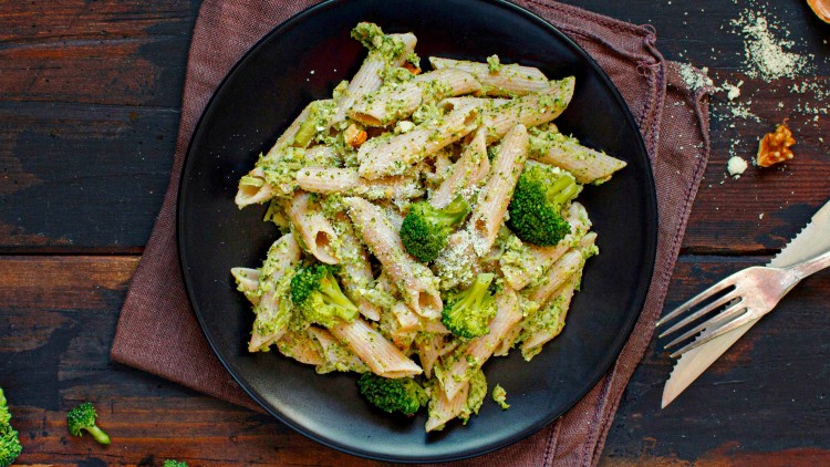 Vollkorn-Penne mit grüner Brokkoli-Pesto-Sauce und geriebenem Käse auf einem schwarzen Teller mit brauner Stoffserviette.