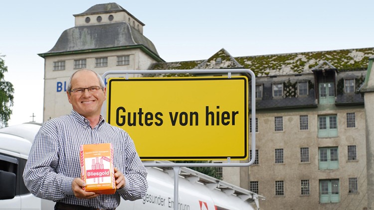 Person hält eine Packung „Bliesgold“ vor einer gelben Tafel mit der Aufschrift „Gutes von hier“, im Hintergrund Gebäude und Lkw.