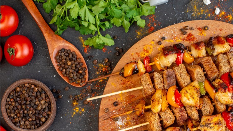 Tomaten, Kräuter, Pfefferkörner und Gewürze auf schwarzem Untergrund, daneben Holzspieße mit Gemüse, Brot und Ananas.
