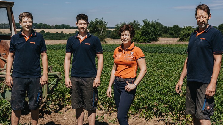 Vier Personen in Arbeitskleidung stehen auf einem Feld mit grünen Pflanzen, im Hintergrund sind Bäume und Felder zu sehen.