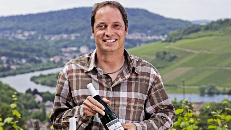 Person in kariertem Hemd hält zwei Weinflaschen vor einer Landschaft mit Fluss, Hügeln und Weinbergen.