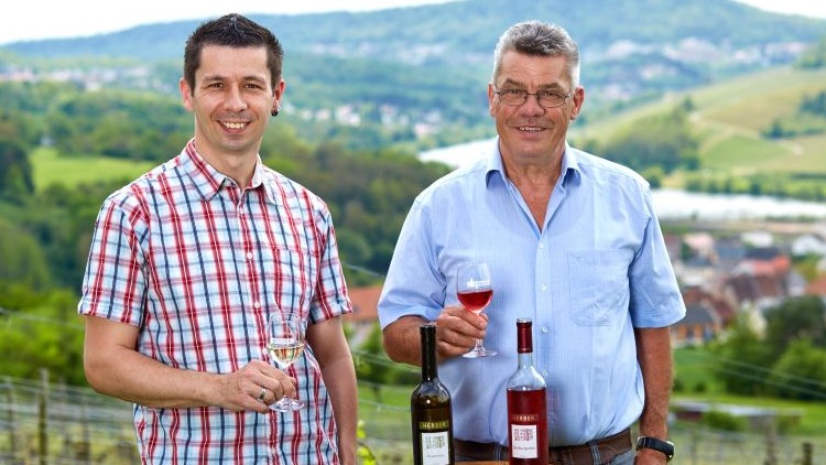 Zwei Personen halten Weingläser mit Weiß- und Roséwein, vor ihnen stehen zwei Weinflaschen auf einem Weinfass, Landschaft im Hintergrund.