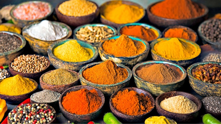 Verschiedene bunte Gewürze in Holzschalen, darunter rote, gelbe und orangene Pulver und ganze Gewürzkörner.