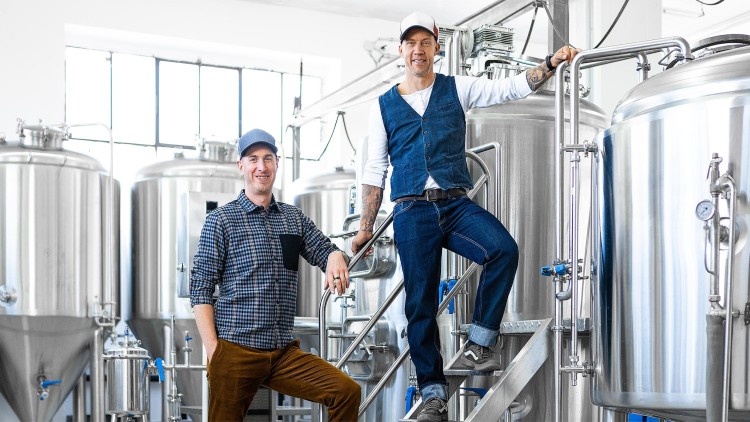 Zwei Personen mit Kappen stehen vor silbernen Braukesseln in einer hellen Brauerei.