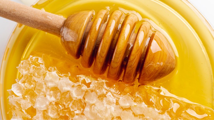 Ein hölzerner Honiglöffel liegt in goldgelbem flüssigem Honig neben einem Stück wabenartigem Honig.