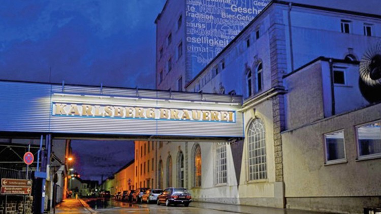 Der beleuchtete Schriftzug „KARLSBERG BRAUEREI“ hängt an einer Überführung zwischen Gebäuden, daneben geparkte Autos bei Nacht.