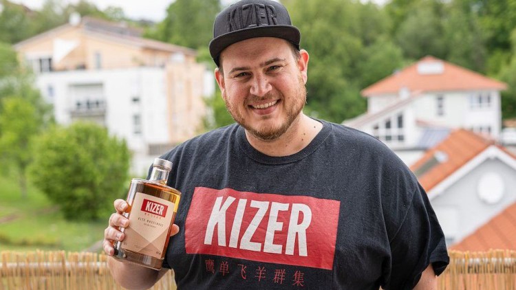 Person hält eine Flasche mit dem Etikett „KIZER Wild Amarillo Spritz“ vor einer unscharfen Häuser- und Grünflächenkulisse.