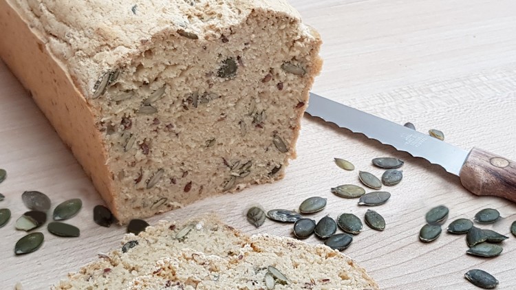 Ein Laib helles Körnerbrot mit Kürbiskernen, daneben liegen ein Brotmesser und lose Kürbiskerne auf einer Holzoberfläche.
