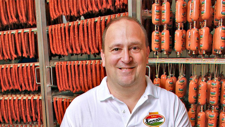 Rote Würste und orange Wurstwaren hängen in mehreren Reihen in einem Kühlraum, Mann trägt ein weißes Hemd mit Kunzler-Logo.