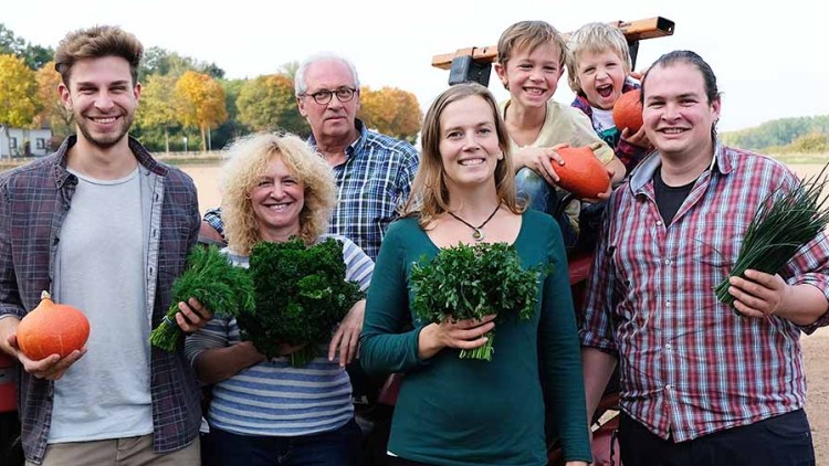 Personen stehen auf einem Feld vor einem Traktor und halten Gemüse wie Kürbis, Kräuter und Schnittlauch in den Händen.