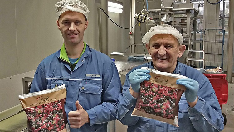 Zwei Personen in blauer Arbeitskleidung halten gefrorene Beerenverpackungen mit der Aufschrift „PAULI GENUSSMANUFAKTUR“.