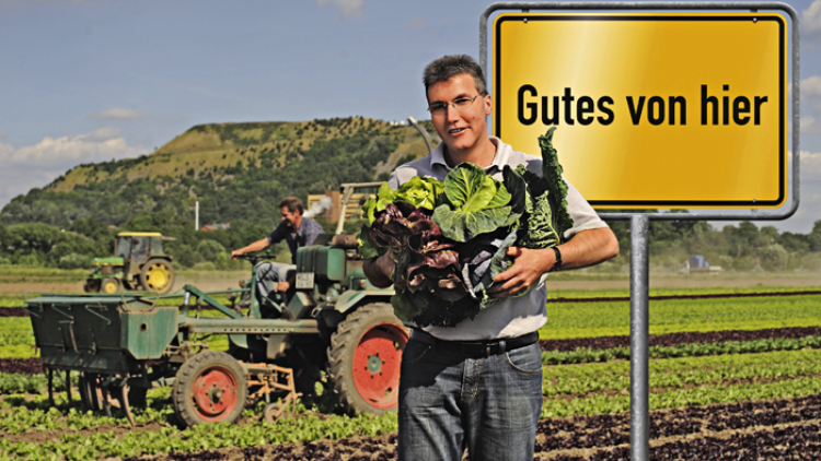 Person hält frisches Gemüse auf einem Feld, im Hintergrund ein Traktor und ein gelbes Schild mit der Aufschrift Gutes von hier.