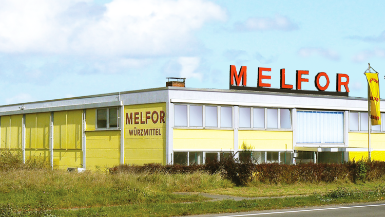 Gelbes Gebäude mit rotem Schriftzug MELFOR und WÜRZMITTEL an der Fassade, drei gelbe Fahnen wehen daneben.