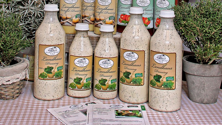 Fünf Flaschen Landhaus-Dressing in unterschiedlichen Größen stehen auf kariertem Tuch vor Kräutertöpfen, Infokarten liegen davor.