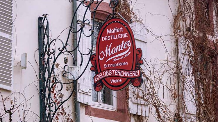 Rotes, ovales Schild mit der Aufschrift „Maffert Destillerie Monter Schnapssideen Edelobstbrennerei Weine“, an Hausfassade.