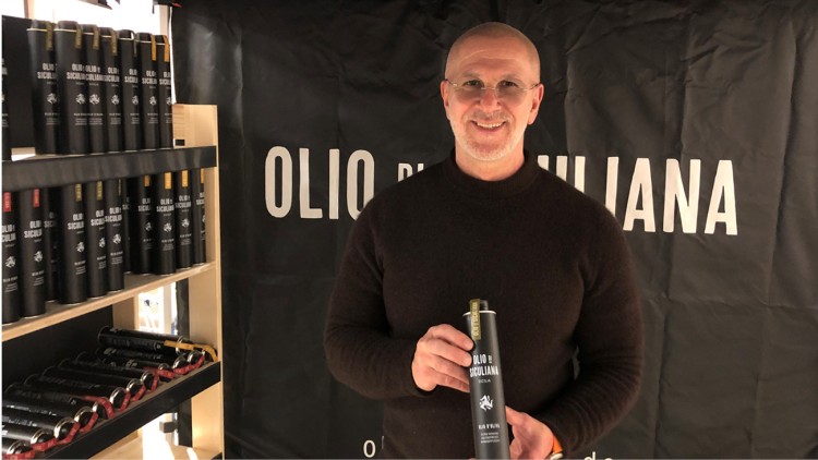 Person hält eine schwarze Ölflasche mit der Aufschrift „Olio di Siculiana“ vor einem schwarzen Banner mit weißer Schrift.