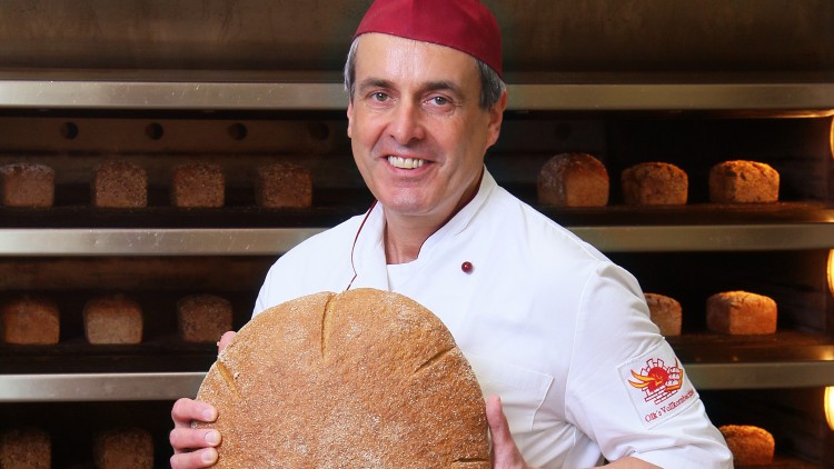 Person in weißer Bäckerbekleidung hält ein großes, rundes Brot vor einem Tisch mit verschiedenem Gebäck, hinten ein Regal mit Broten.